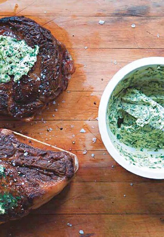 Two steaks on a cutting board topped with herbed compound butter an a bowl of the butter on the side.