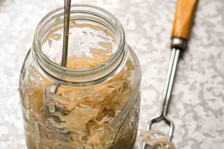 A glass jar of single quart sauerkraut with a spoon inside and a two-pronged fork lying beside it.