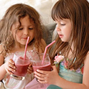Two young girls sitting and holding berry smoothies with straws.