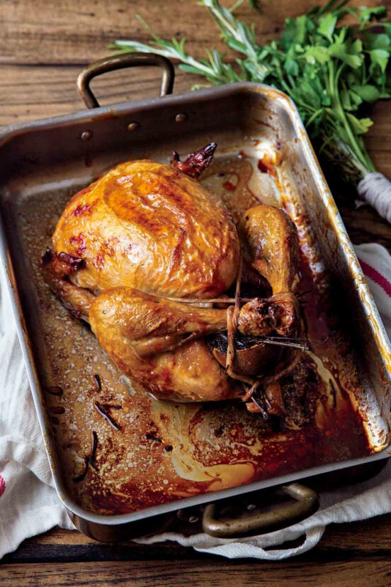 A perfect roast chicken in a metal roasting pan that is resting on a kitchen towel, with a bunch of herbs beside it.