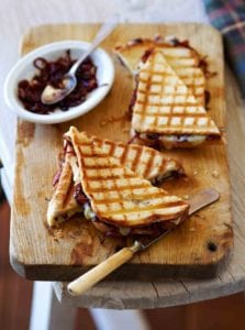 Two halved grilled cheese with onion jam on a wooden cutting board alongside a dish of onion jam and a butter knife.