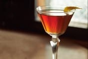 A small aperitif glass filled with classic Manhattan cocktail and a lemon twist garnish.