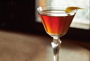 A small aperitif glass filled with classic Manhattan cocktail and a lemon twist garnish.