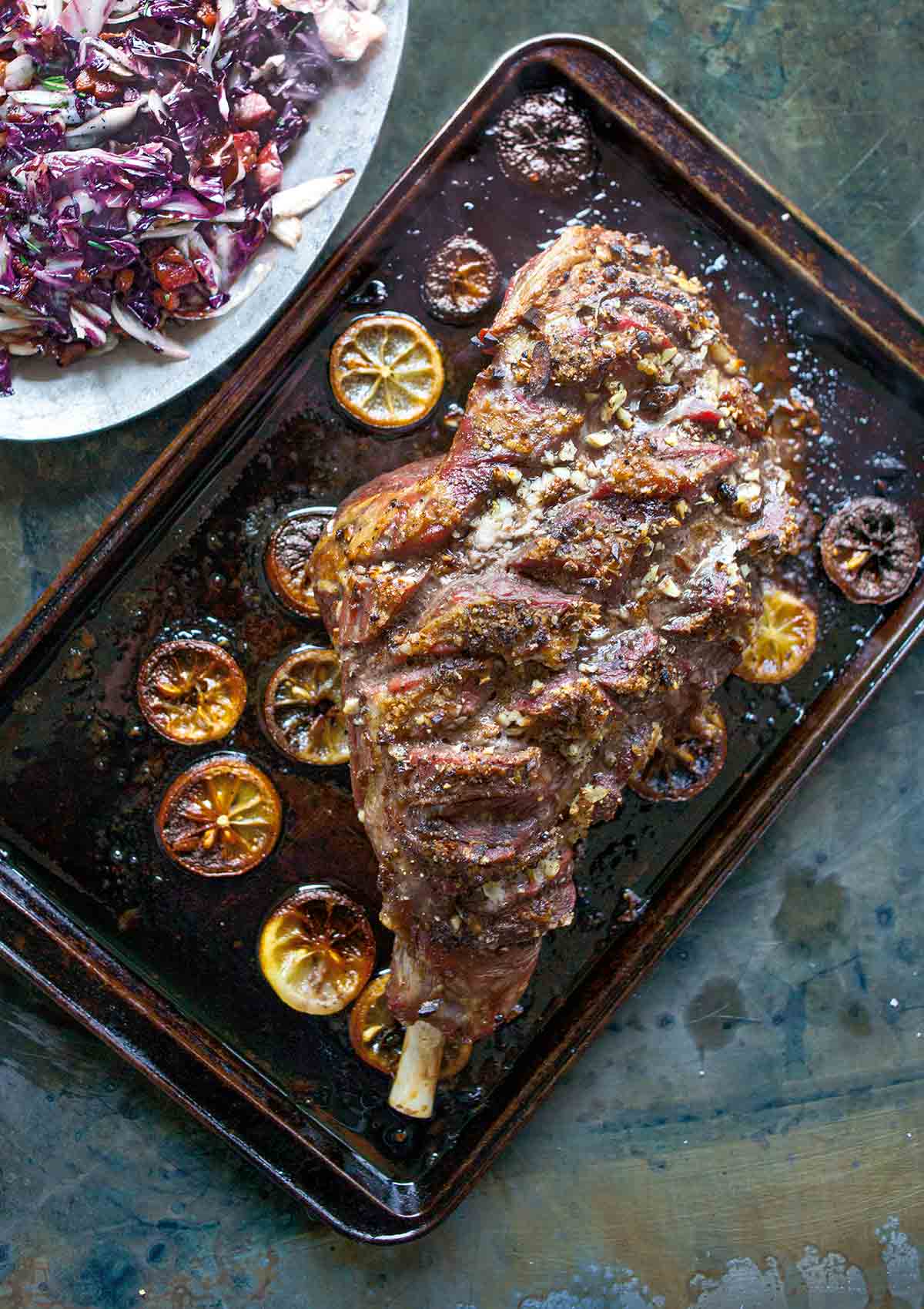 A whole roast leg of lamb on a rimmed baking sheet with charred lemon slices surrounding the lamb.