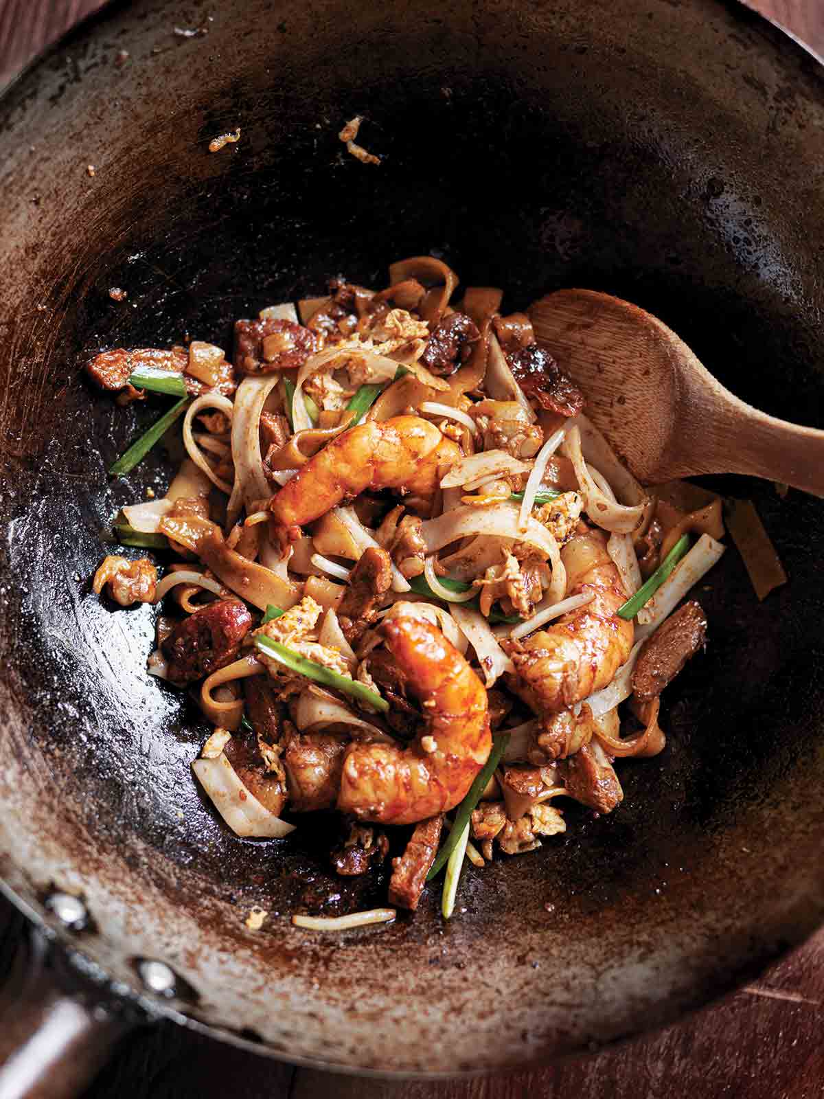 A metal wok with a serving of char kway teow and a wooden spoon resting inside.