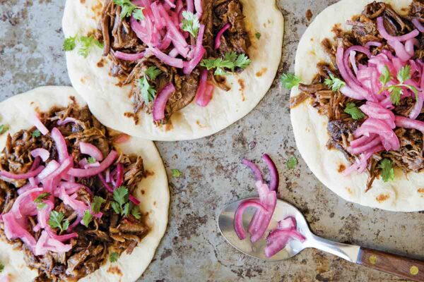 Three ancho short rib tacos on flour tortillas, topped with pickled red onion and cilantro.