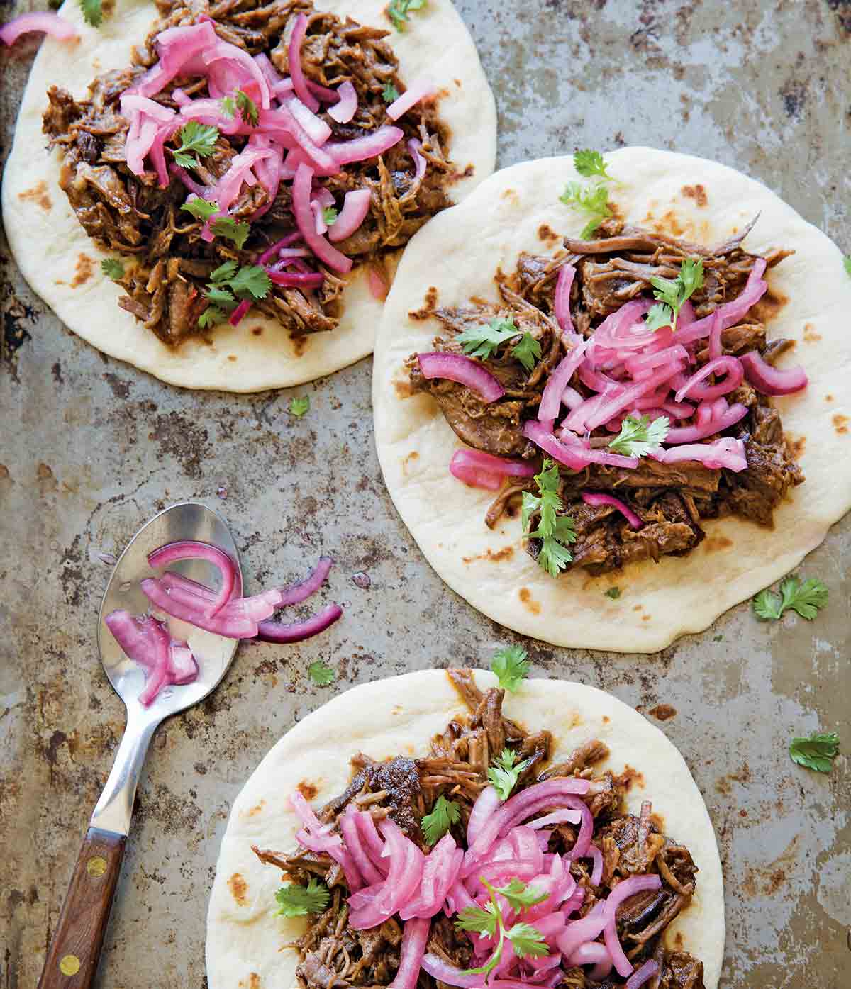 Three ancho short rib tacos on flour tortillas, topped with pickled red onion and cilantro.