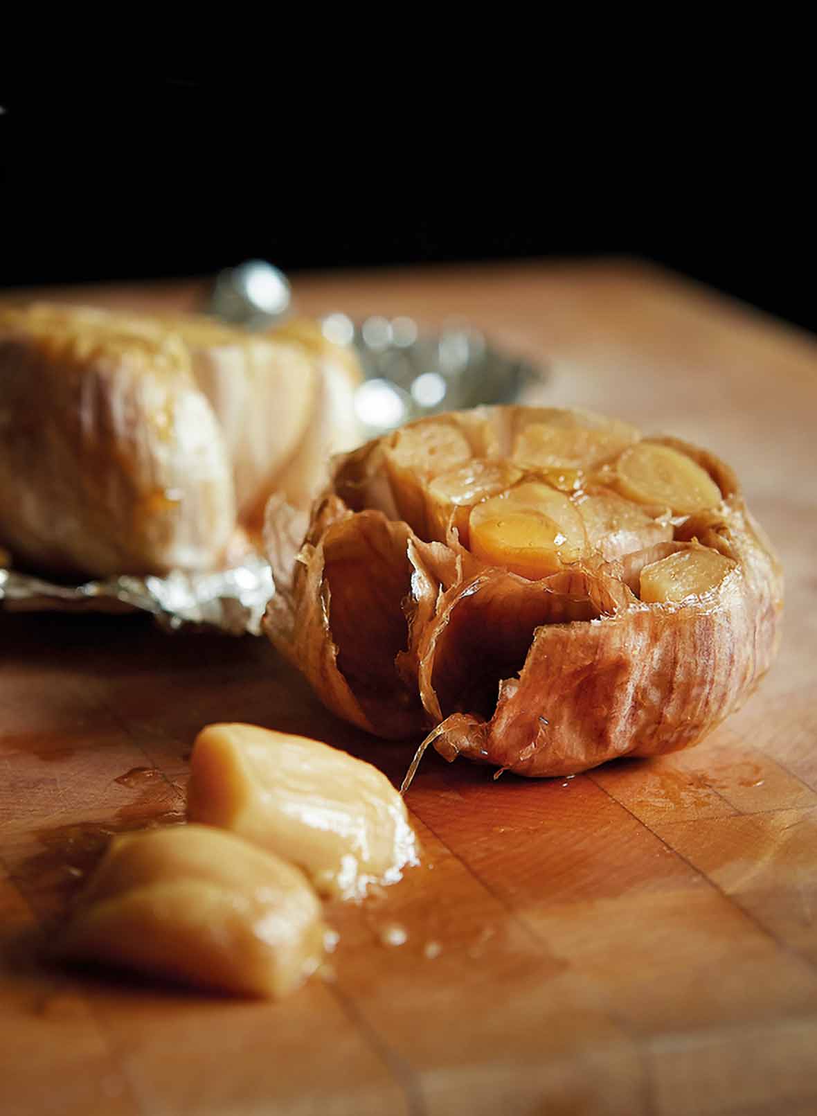 Two heads of roasted garlic--one in foil, one unwrapped--drizzled with oil on a cutting board.