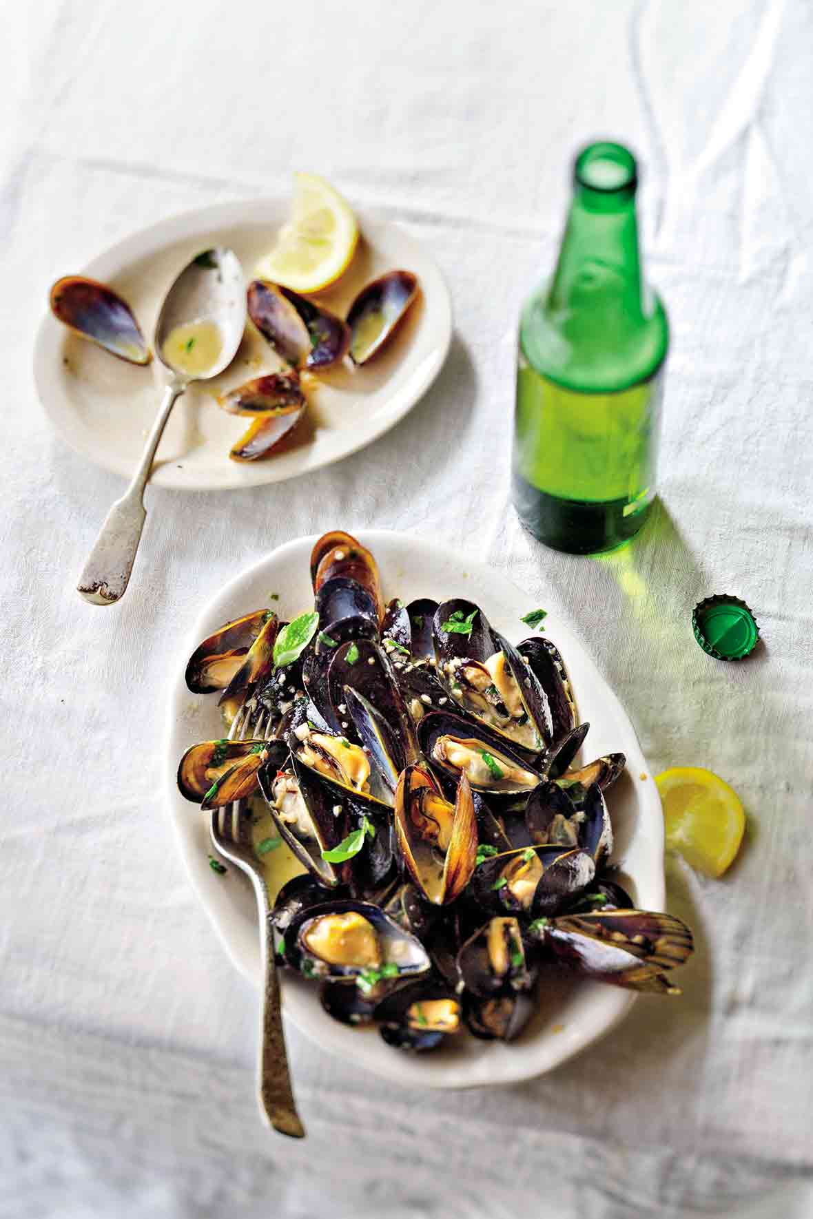 White plate with steamed mussels in beer, topped with parsley, nearby lemon wedges and a bottle of beer