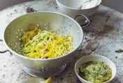 A wooden table with a colander holding veggie spaghetti, a small white bowl of pesto, and a small dish of lemon wedges.