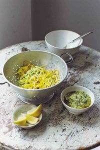 A wooden table with a colander holding veggie spaghetti, a small white bowl of pesto, and a small dish of lemon wedges.