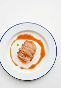 A sliced duck breast with maple bourbon sauce drizzled around it on a blue-rimmed white plate.