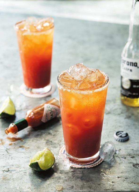 Two glasses filled with Mexican blood Mary, some lime wedges, a mostly empty bottle of Corona, and a bottle of Tabsco on its side.