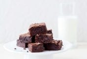 A white cake stand of a pile of one-pot cocoa brownies, a glass of milk, on a white table