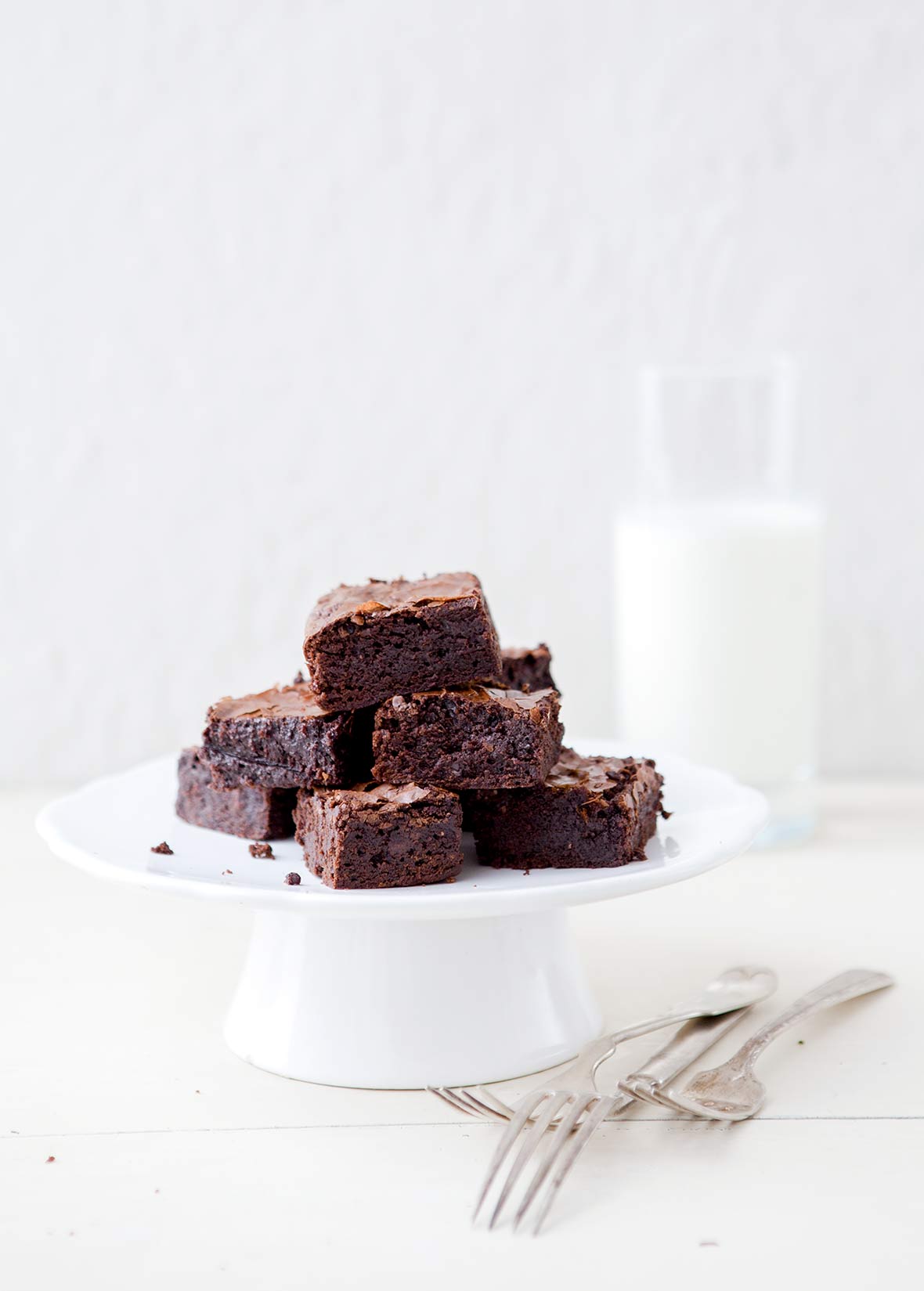 A white cake stand of a pile of one-pot cocoa brownies, a glass of milk, on a white table