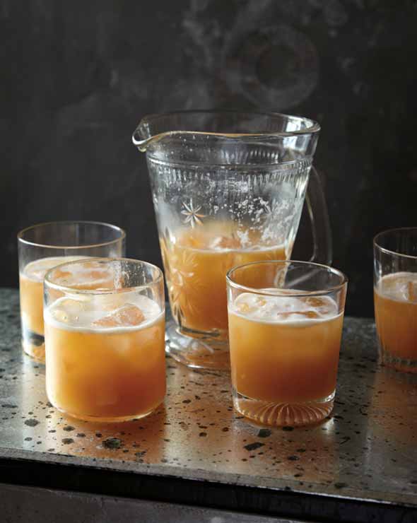 A pitcher and 4 tumblers filled with amber colored whiskey sour.