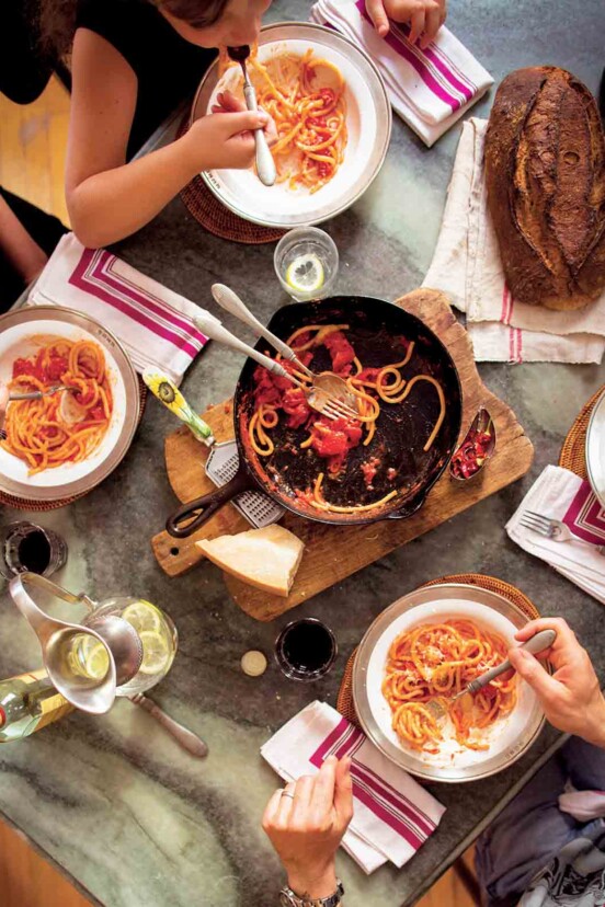 People gathered around a cast-iron skillet on a wooden board with some spaghetti and red sauce.