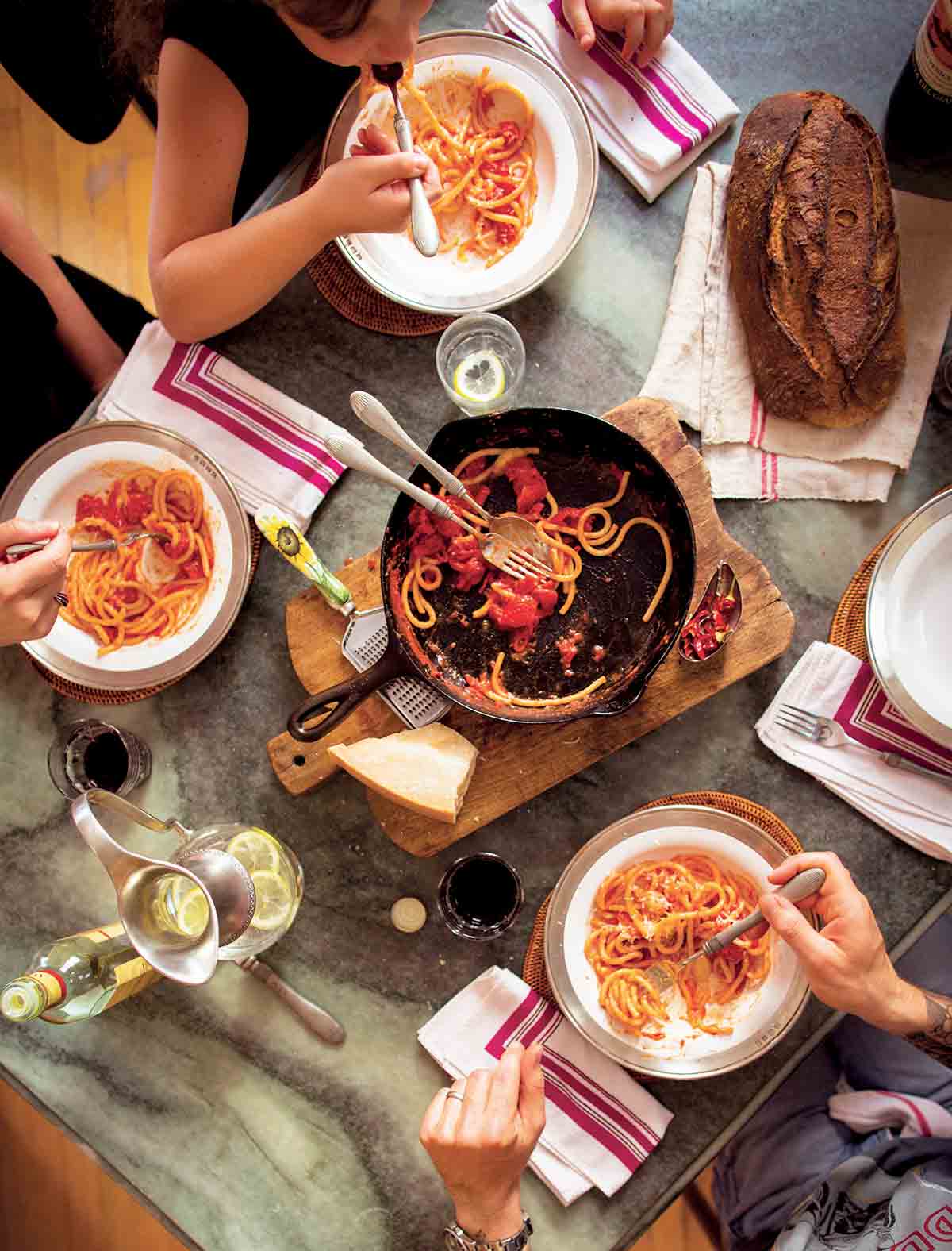 People gathered around a cast-iron skillet on a wooden board with some spaghetti and red sauce.