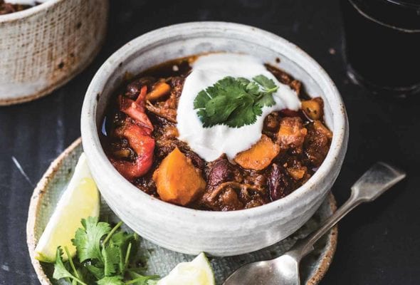A bowl of shredded beef chili with sweet potatoes on a plate containing a spoon, two lime wedges, and cilantro leaves.