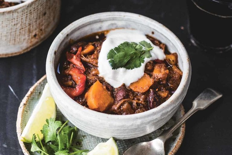 A bowl of shredded beef chili with sweet potatoes on a plate containing a spoon, two lime wedges, and cilantro leaves.