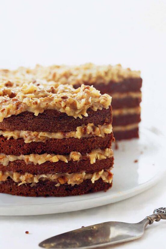 A four-layer German chocolate cake with a slice taken out on a white plate with a cake server nearby.