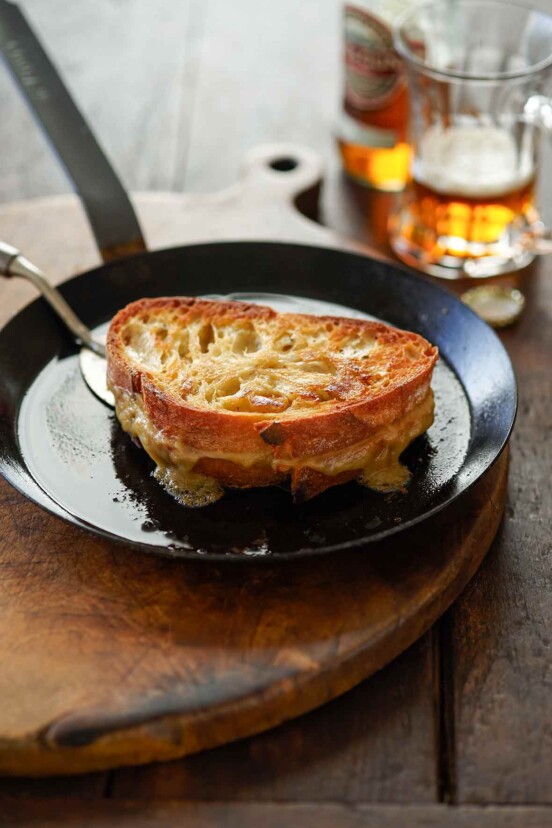 A black metal skillet with a spatula ready to flip a Welsh rarebit grilled cheese sandwich.