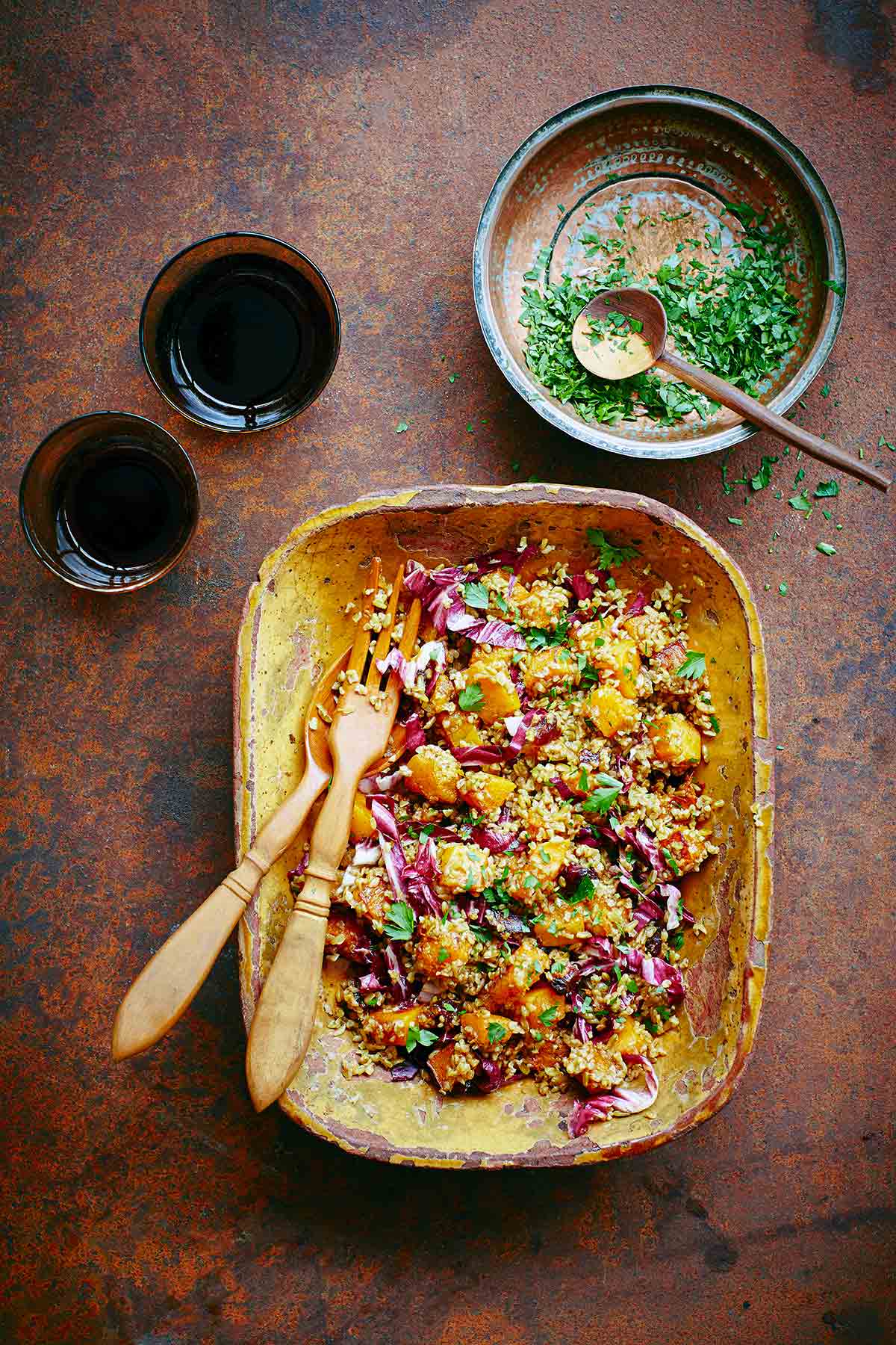 A wooden bowl with butternut squash and whole grain salad with a bowl of chopped parsley on the side and two glasses.