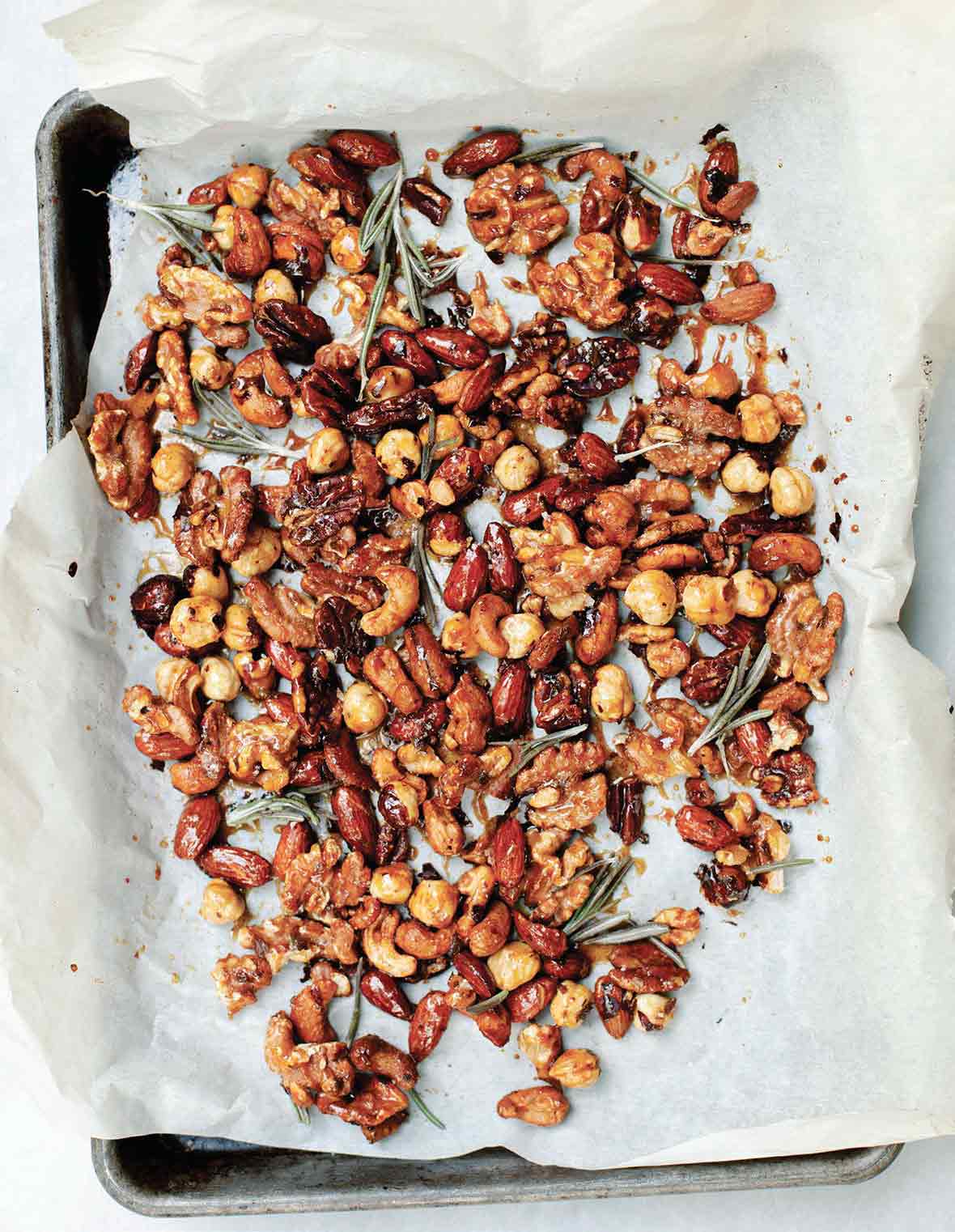 A layer of assorted nuts and rosemary sprigs on a sheet of parchment paper resting in a sheet pan