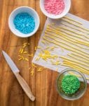 Parchment paper with long lines of yellow homemade sprinkles, plus bowls of green, blue, and pink sprinkles
