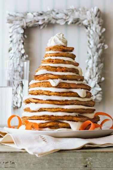 A stack of carrot cake pancakes layered with mascarpone frosting, shaped to look like a Christmas tree on a white plate.
