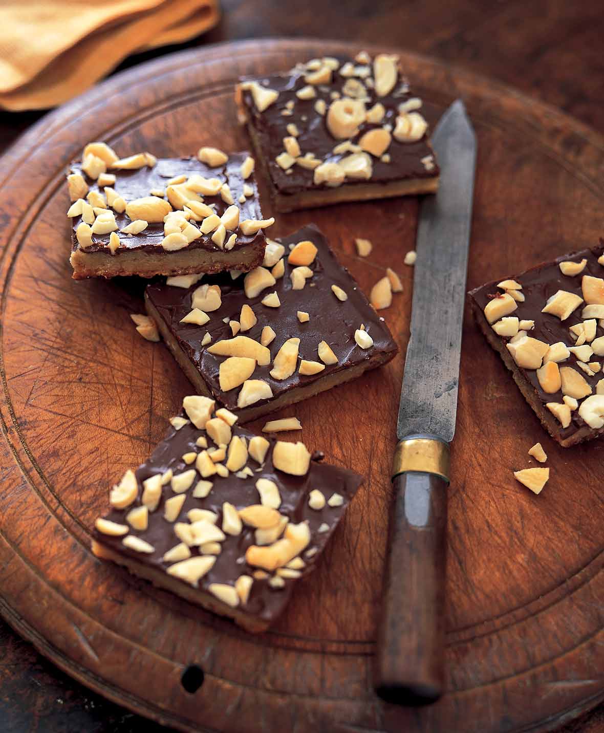 Four pieces of cashew caramel cracker bars on a round wooden board with a knife resting beside them.