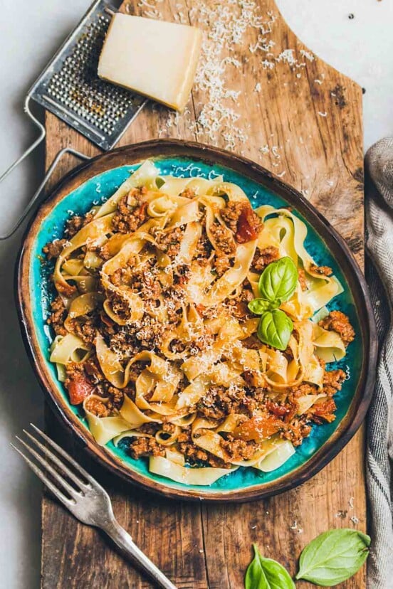 A blue bowl filled with pappardelle noodles and Marcella Hazan's bolognese sauce on a wooden board with a block of Parmesan and a grater beside the bowl.
