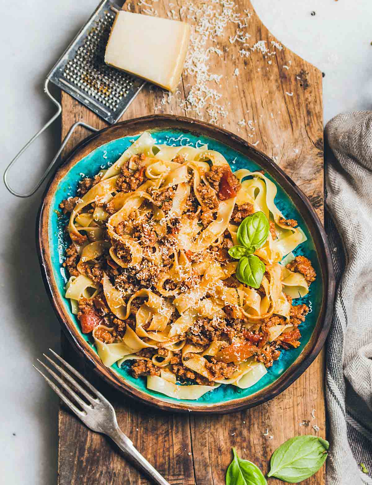 A blue bowl filled with pappardelle noodles and Marcella Hazan's bolognese sauce on a wooden board with a block of Parmesan and a grater beside the bowl.