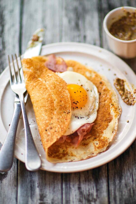 A thin pancake filled with ham and an egg on a white plate, along with a fork and knife.