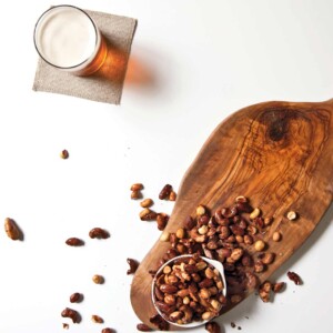 An overflowing bowl of beer nuts on a wooden board with a glass of beer on a napkin beside it.