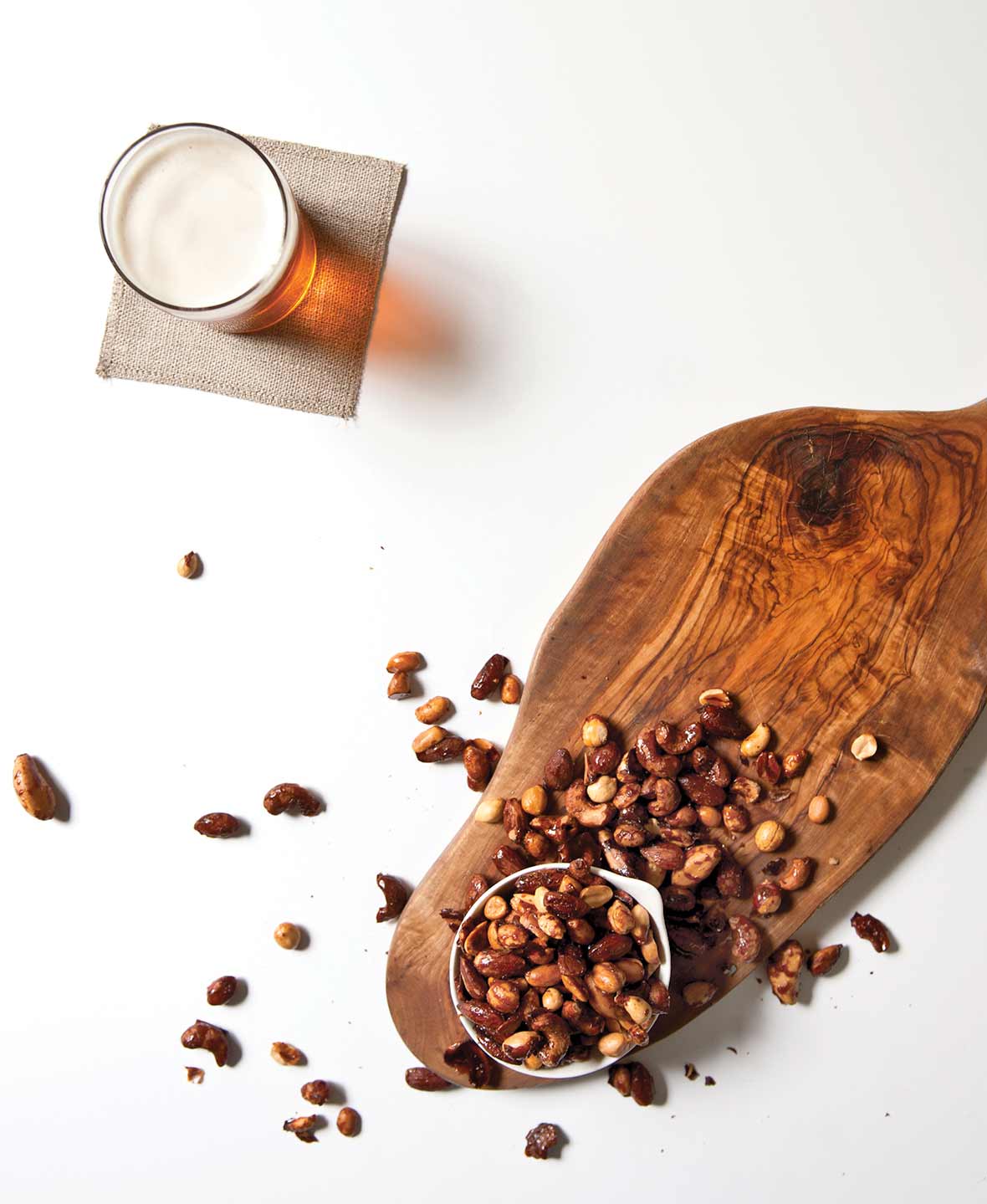 An overflowing bowl of beer nuts on a wooden board with a glass of beer on a napkin beside it.