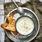 A bowl of cauliflower soup sitting on a metal pie plate with a spoon and slices of toasted baguette