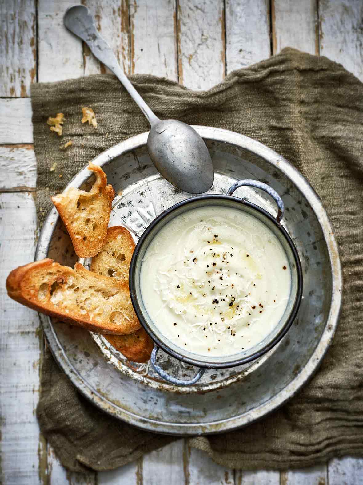 A bowl of cauliflower soup sitting on a metal pie plate with a spoon and slices of toasted baguette