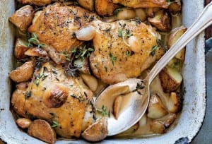 A baking dish with chicken thighs topped with garlic cloves, red potatoes, and thyme. A silver spoon is nestled in the dish.