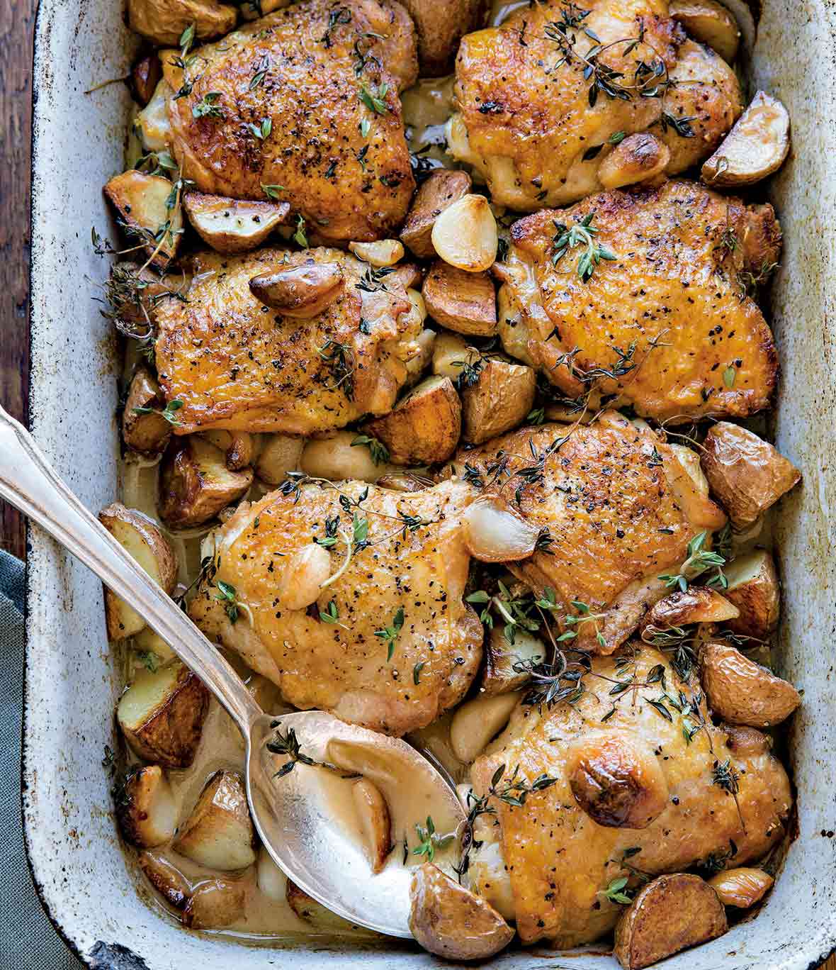 Cheap cuts: A baking dish with chicken thighs topped with garlic cloves, red potatoes, and thyme. A silver spoon is nestled in the dish.