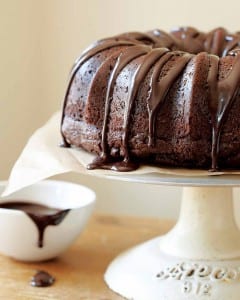 A chocolate glazed chocolate bundt cake sitting atop a white cake stand with a dish of glaze on the side