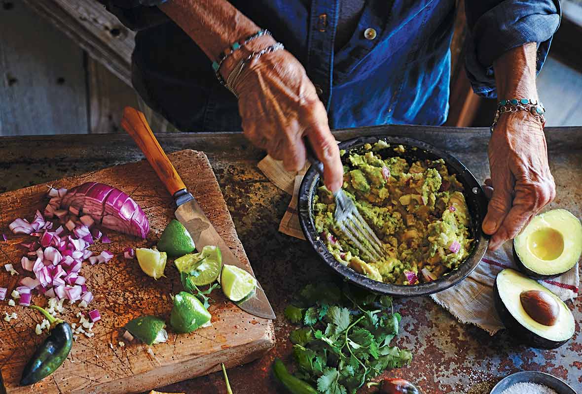 A person mashing avocado into a bowl of easy guacamole on a table with salt, avocado, cilantro, lime, and chiles.