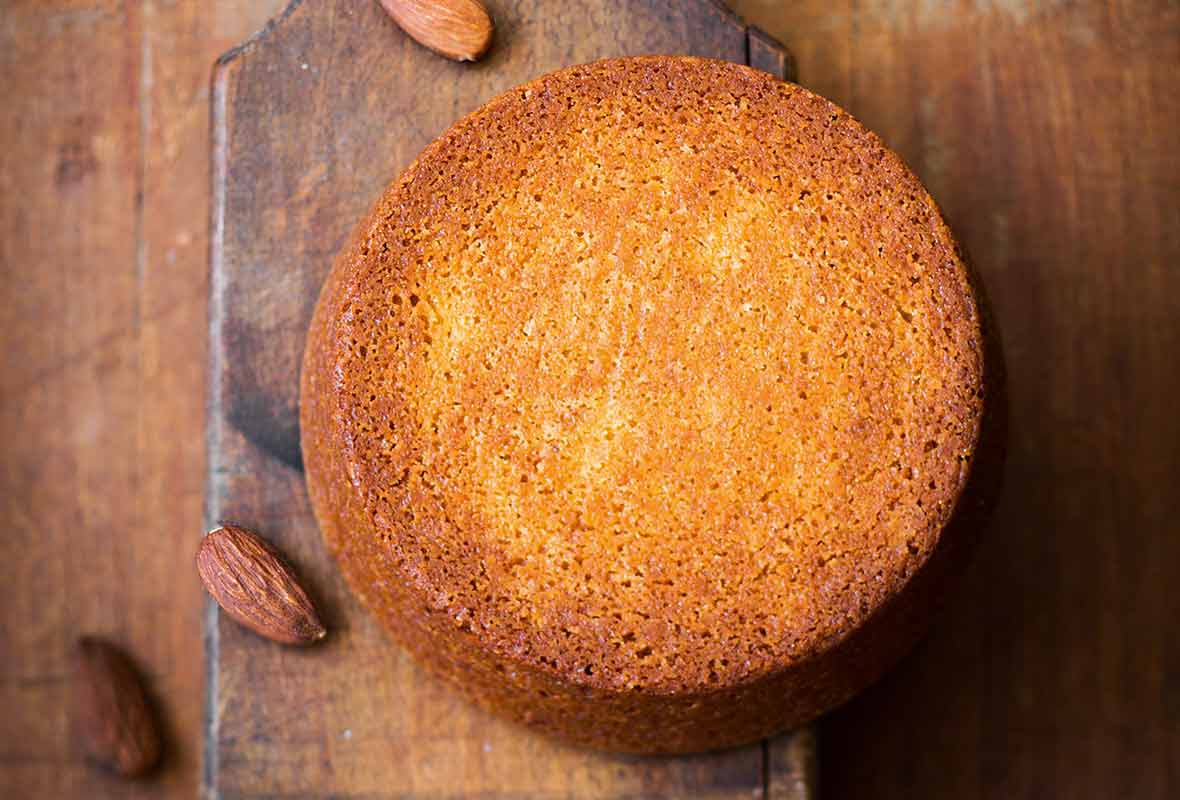 A round flourless almond cake on a wooden baking paddle with almonds scattered around it