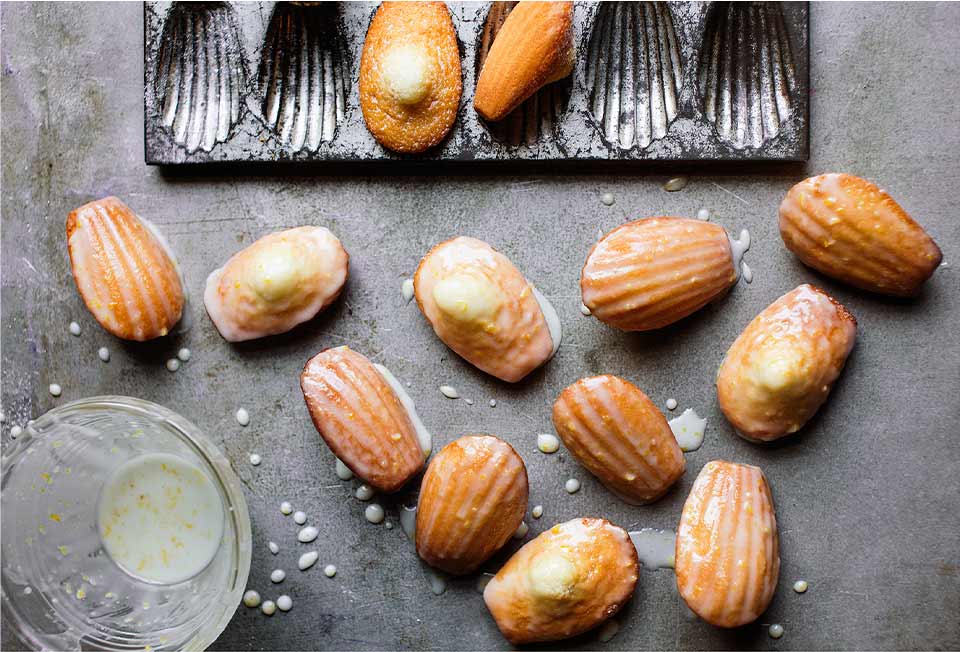 Several glazed lemon madeleines scattered next to a madeleine pan and a dish of glaze.