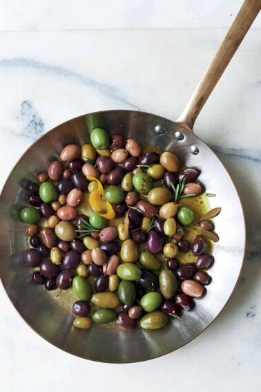 A metal skillet filled with warm olives, rosemary, and lemon zest.