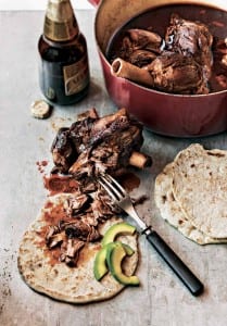 A beer braised lamb shank in a red Dutch oven, and another shank on a surface next to tortillas, sliced avocado, and an open beer.