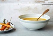 A white bowl filled with carrot ginger soup, with a spoon resting inside and a plate of carrots beside it.