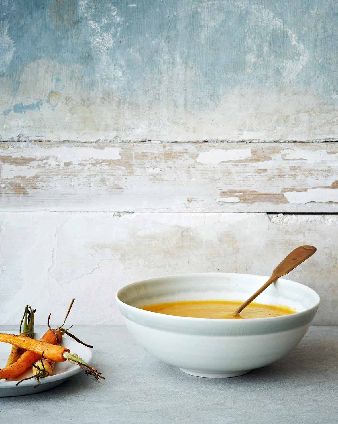 A white bowl filled with carrot ginger soup, with a spoon resting inside and a plate of carrots beside it.