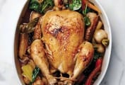 A whole chicken in a pot surrounded by vegetables including carrots, baby turnips, and spinach.