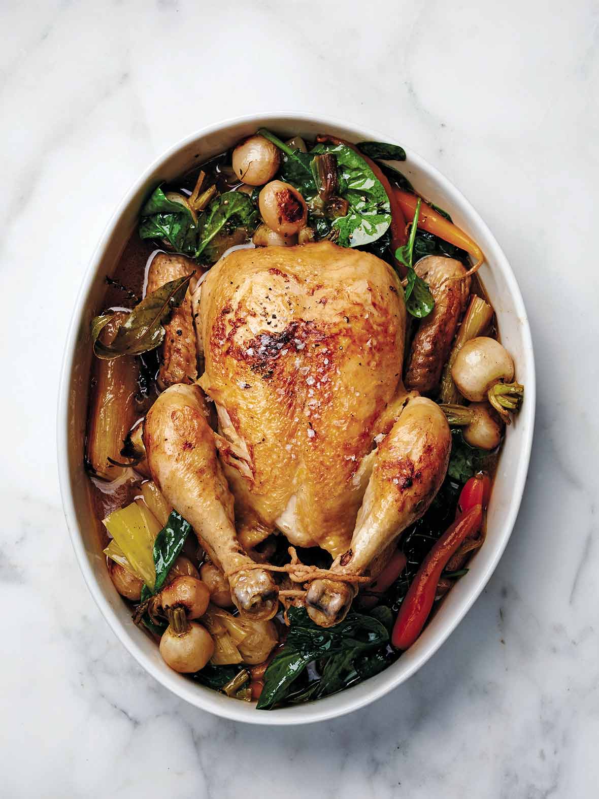 A whole chicken in a pot surrounded by vegetables including carrots, baby turnips, and spinach.
